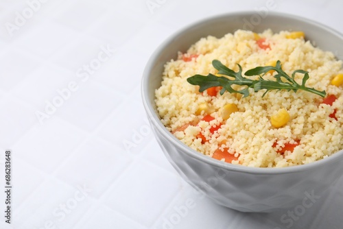 Tasty couscous with pepper, corn and arugula in bowl on white tiled table, closeup. Space for text