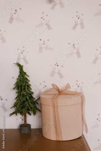Small Christmas tree souvenir and a round gift box with a ribbon and a bow on a shelf in the interior of a cozy house