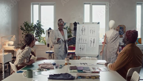 Full shot of diverse atelier workers sitting at table and business owner coming with tablet and speaking about new apparel collection using whiteboard with sketches in tailoring studio photo