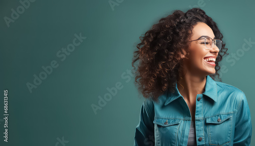 woman in denim jacket and curly hair with glasses laughing on blue background