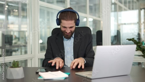 Smiling young worker in wireless headphones pretending to playing invisible piano at personal workplace. Energetic caucasian businessman having break after working on modern laptop in office.