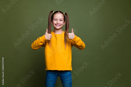 Photo of optimistic smart schoolgirl with ponytails wear yellow pullover two hands show thumbs up isolated on khaki color background