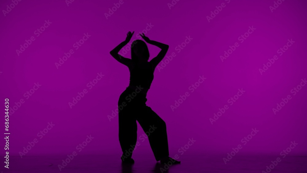In the frame on the purple background in the silhouette. Dances slender, beautiful girl. Demonstrates dance moves in the style of hip hop, lifting hands up. It is feminine, plastic, rhythmic