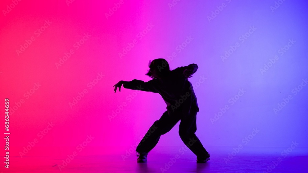 Fototapeta premium In the frame on the purple, pink, gradient background in the silhouette. Dances slim, beautiful girl. Shows dance moves in the style of hip hop, hands to the side. It is feminine, plastic, rhythmic