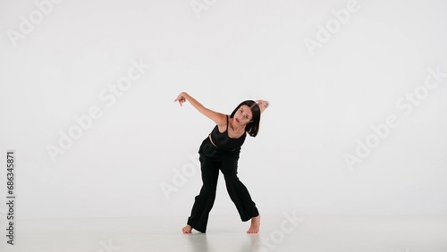 In the frame on a white background. A young, beautiful girl dances. Demonstrates dance moves in the style of hip hop. She gazes at the camera. Shes feminine, barefoot in a black top and pants