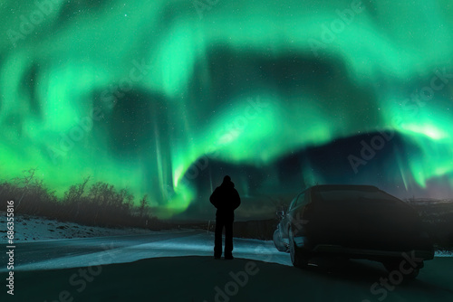 Silhouette of unrecognizable man and dark car on mountain road in front of Northern green lights shine over mountains in Sweden, Lapland. Night photo, Aurora, rear side photo photo