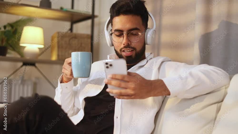 Attractive young man with cup of hot coffee or tea while hold smartphone checking email watching social media indoors Smiling bearded guy with headphones listening to music texting on phone at home Attractive young man with cup of hot coffee or tea while hold smartphone checking email watching social media indoors Smiling bearded guy with headphones listening to music texting on phone at home