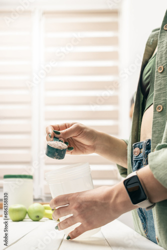 Young woman in jeans and shirt with measuring spoon in her hand puts portion of whey protein powder into shaker on white wooden table with bananas and apples. Process of making protein drink photo