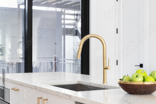 A gold kitchen faucet detail with a white marble countertop and island  a bowl of green apples  and a stainless steel sink.