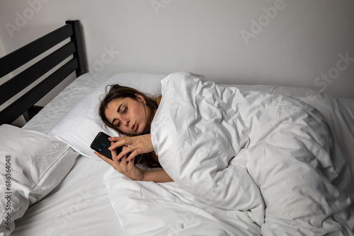 A girl in bed with a phone on a white bed in a dark room.