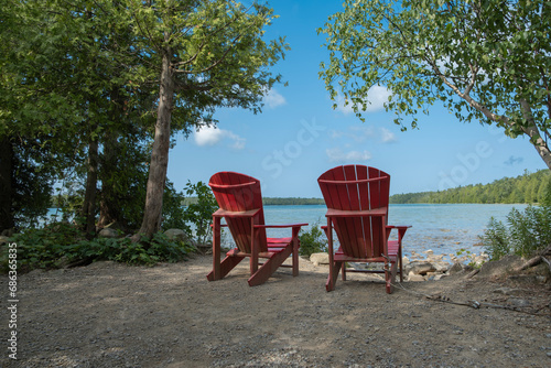 Two bright red wooden muskoka adirondack chairs on the Georgian Bay shore in Brice Peninsula National Park surrounded by green trees, shot on a sunny summer day. photo