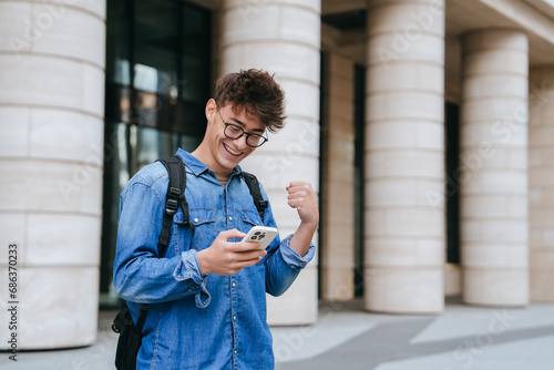 Excited caucasian student guy in jeans shirt holds phone reads message clenches fist smiles wide. Success concept. Teenager celebrating win, passed exam. Happy people. photo