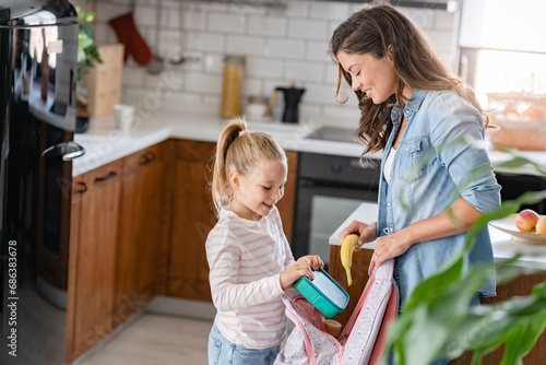 The cute girl is standing next to her caring young mother, who is preparing her to go to school giving her a fruit snack in a lunch box photo