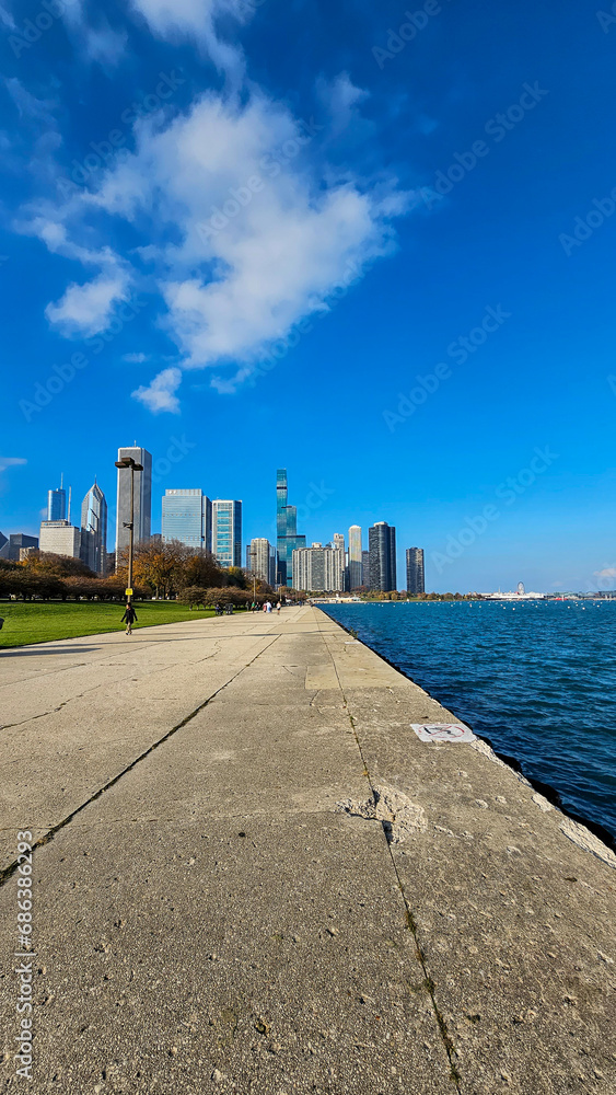 A beautiful autumn landscape at Lakefront Park with the rippling blue waters of Lake Michigan, autumn trees, people walking, skyscrapers and office buildings, blue sky and clouds in Chicago Illinois