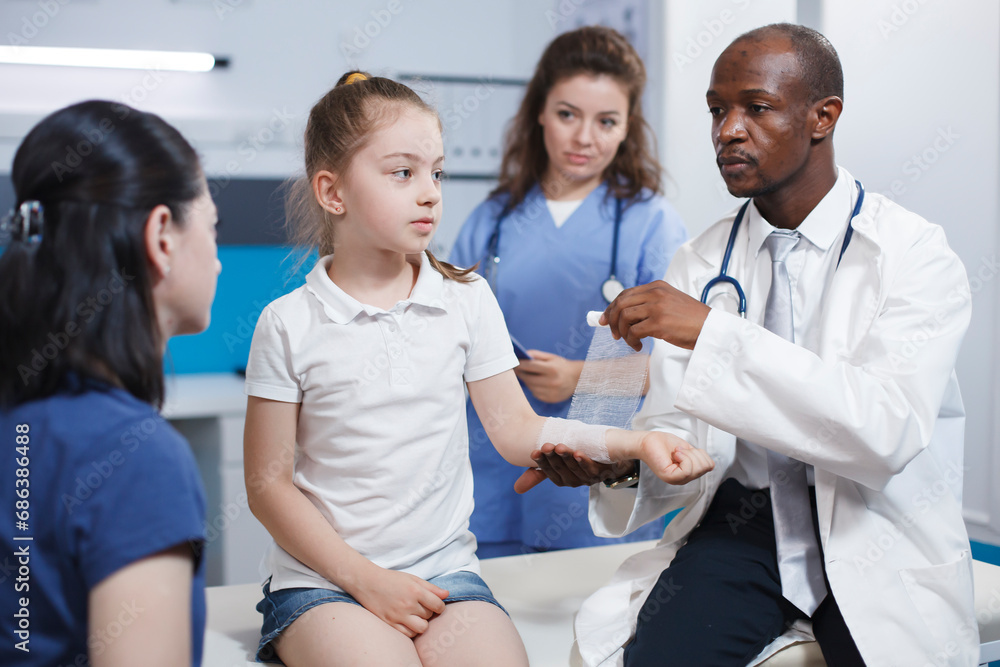 Obraz premium Arm of girl patient being meticulously wrapped with a sterile bandage by multicultural medical personnel. Caucasian youngster accompanied by her mother is being treated by doctor and nurse.