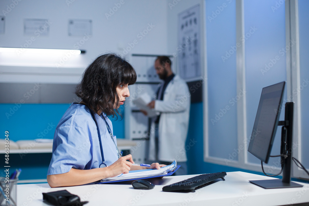 Professional medic looking at computer screen and medical files for healthcare. Nurse reviewing information on pc monitor and documents for assistance and support, doing overtime work. Professional medic looking at computer screen and medical files for healthcare. Nurse reviewing information on pc monitor and documents for assistance and support, doing overtime work.
