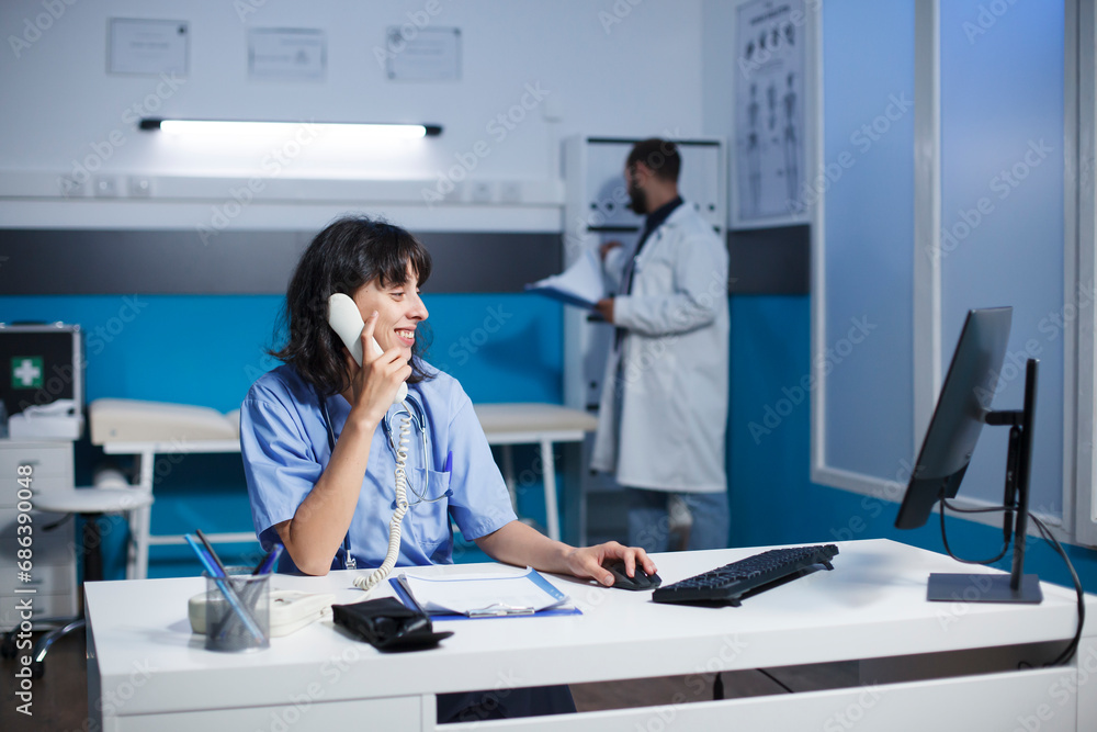 Caucasian doctor and nurse collaborate in a modern hospital office. Engaged in a detailed discussion is a female practitioner holding a landline phone while using a desktop computer.