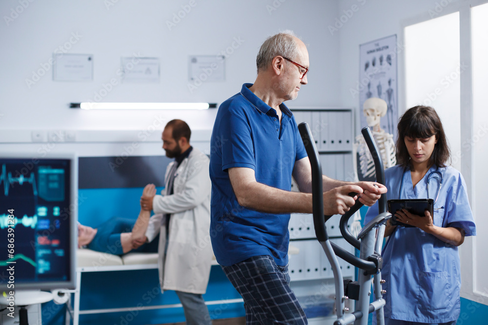 Obraz premium Elderly male patient is engaged in physiotherapy on stationary bicycle, with support from nurse in blue scrubs. Medical assistant with digital tablet helping senior man with physical therapy.