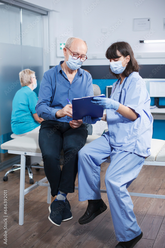 Female medical assistant holding a clipboard with a hospital form for male elderly patient to sign. Nurse practitioner and old man seated on clinic bed looking at paperwork, both wearing face masks. Female medical assistant holding a clipboard with a hospital form for male elderly patient to sign. Nurse practitioner and old man seated on clinic bed looking at paperwork, both wearing face masks.