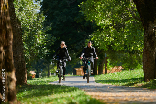 A blissful couple, adorned in professional cycling gear, enjoys a romantic bicycle ride through a park, surrounded by modern natural attractions, radiating love and happiness