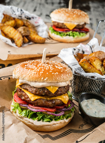 Beef burger and double burger with lettuce, tomatoes, slice of cheese, pickles, sauce, onions rings, french fries on craft paper over wooden background, close up