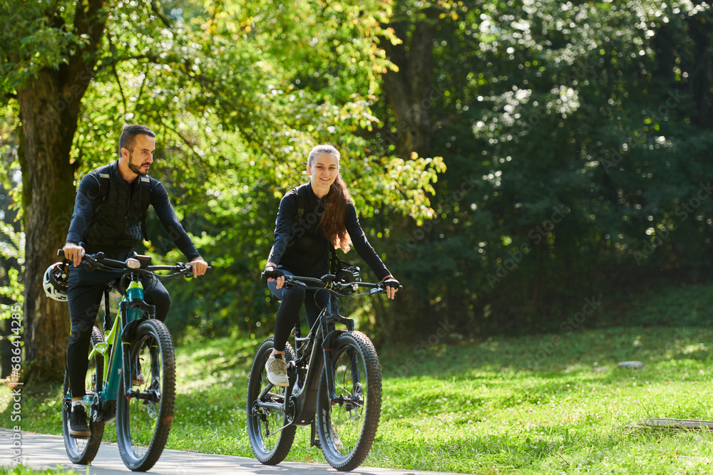 Naklejka premium A blissful couple, adorned in professional cycling gear, enjoys a romantic bicycle ride through a park, surrounded by modern natural attractions, radiating love and happiness