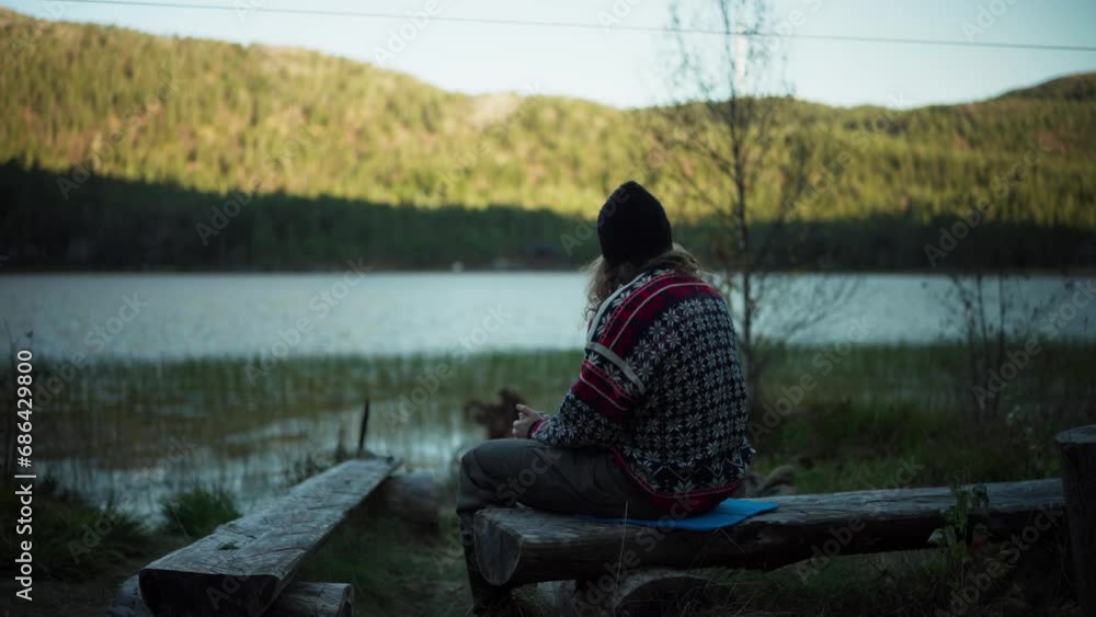Man Sitting On Tree Trunk Bench While Drinking Coffee By The Lake In The Morning. - static shot Man Sitting On Tree Trunk Bench While Drinking Coffee By The Lake In The Morning. - static shot