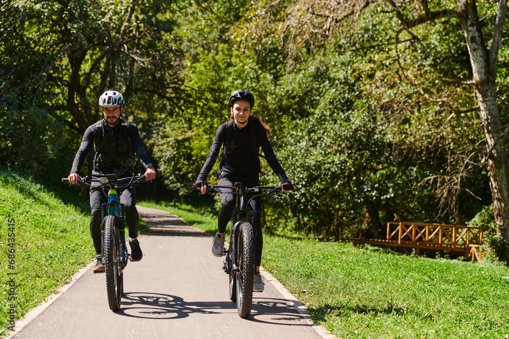 Obraz premium A blissful couple, adorned in professional cycling gear, enjoys a romantic bicycle ride through a park, surrounded by modern natural attractions, radiating love and happiness