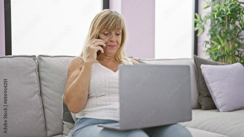 Relaxed middle age blonde woman comfortably speaking on her smartphone, concentrating on her laptop talk, while resting at home on the living room sofa Relaxed middle age blonde woman comfortably speaking on her smartphone, concentrating on her laptop talk, while resting at home on the living room sofa