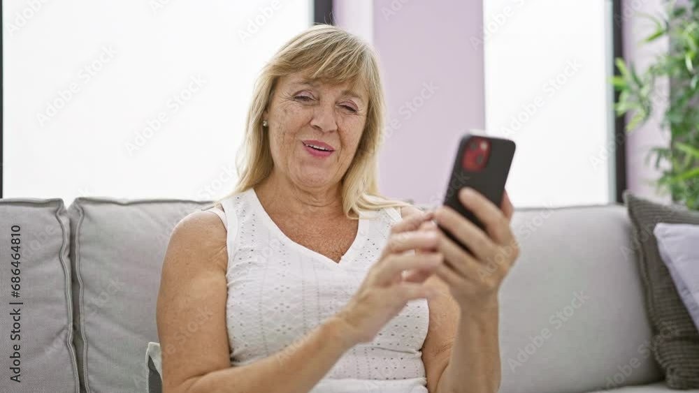 Joyful middle-aged blonde woman enjoying tech-time, smiling while pointing at smartphone, sitting comfortably on living room sofa at home Joyful middle-aged blonde woman enjoying tech-time, smiling while pointing at smartphone, sitting comfortably on living room sofa at home