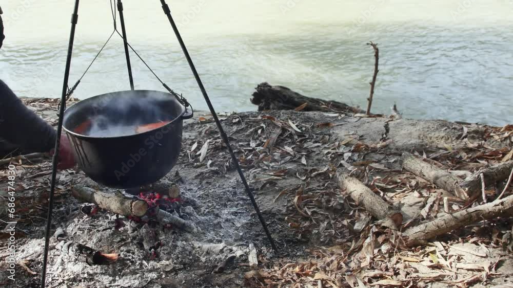 hand adds wood to the fire, cooks fish soup in a cauldron on a fire in nature hand adds wood to the fire, cooks fish soup in a cauldron on a fire in nature