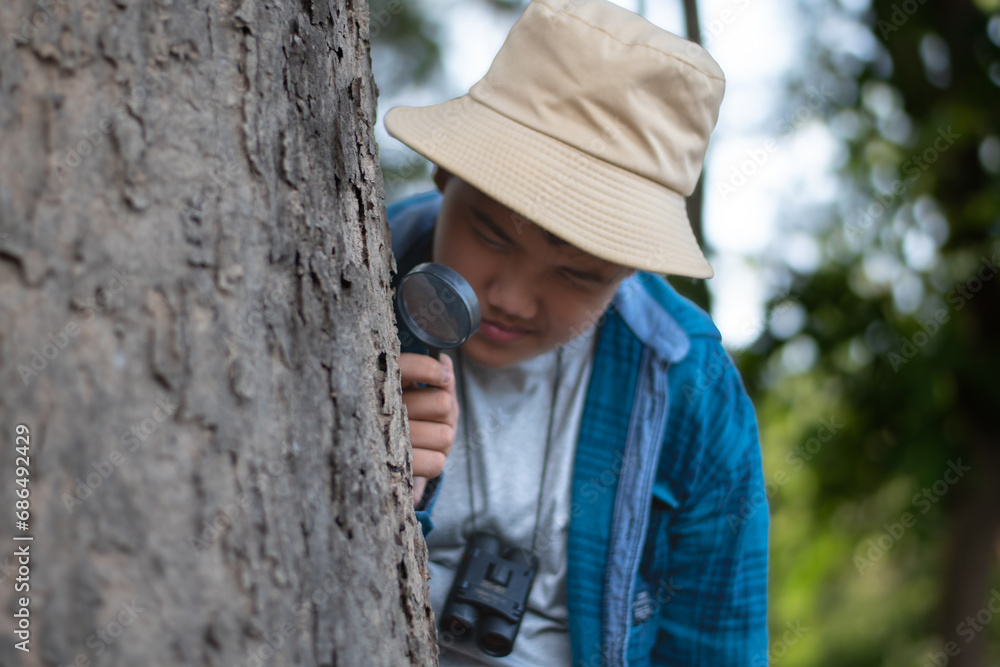 Asian boy using outdoor mini magnifying glass to watch moss pattern which growing on tree trunk during study various plant species for searching in the school's botanical garden library after school.