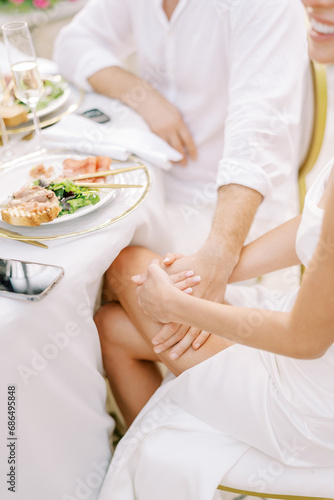 Bride holds groom hand on her knee while sitting at the festive table. Cropped. Faceless