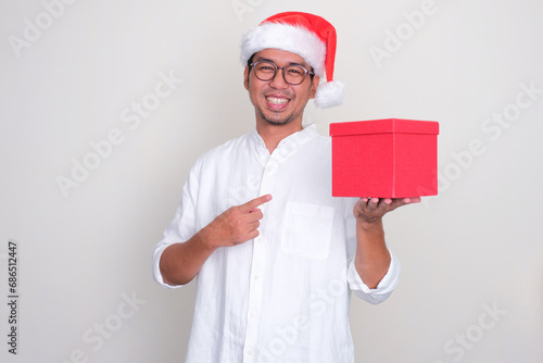A man wearing christmas hat smiling happy while pointing to gift box that he hold photo