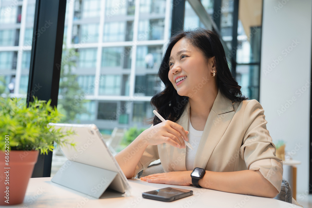 Smiling Asian freelance woman holding pen and working on laptop computer on table at coffee shop Smiling Asian freelance woman holding pen and working on laptop computer on table at coffee shop