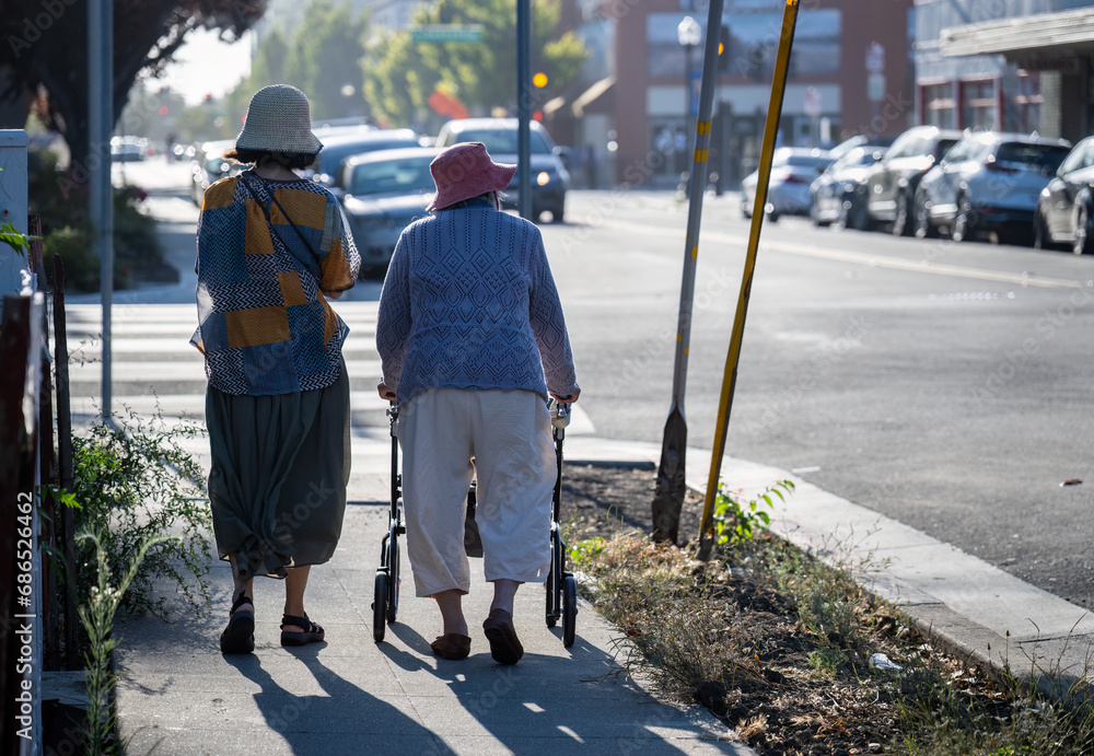 Senior woman walking using a mobility walker on the pedestrian walkway in the city, accompanied by her daughter.