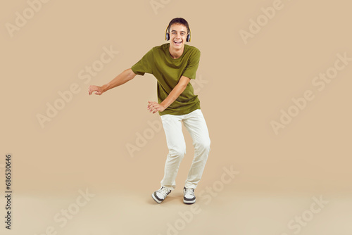 Full length portrait of a young joyful boy wearing casual clothes having fun, dancing and listening to music in headphones, looking at camera isolated on a studio beige background.