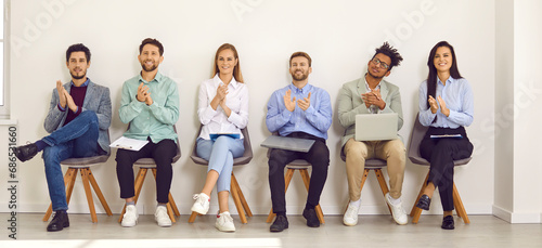 Young professional business people sitting on the chairs in a row looking at the camera and applauding. Job candidates seekers with resumes and laptops or group of a staff on meeting. Banner.