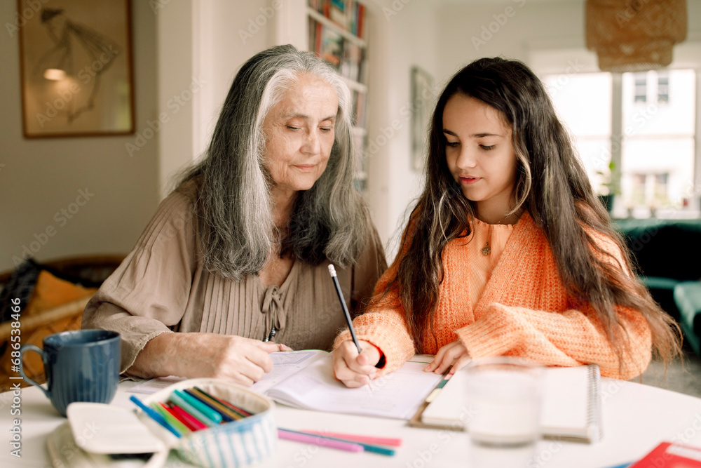 Senior woman assisting granddaughter in homework while sitting at table Senior woman assisting granddaughter in homework while sitting at table