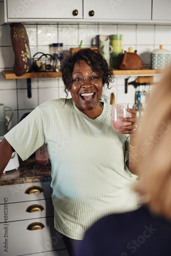 Happy mature woman with smoothie glass talking to female friend at home