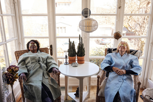 Multiracial female friends relaxing with cucumber slices on eyes at home photo
