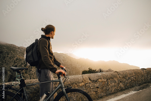 Side view of young man looking at mountains while standing with bicycle on road photo
