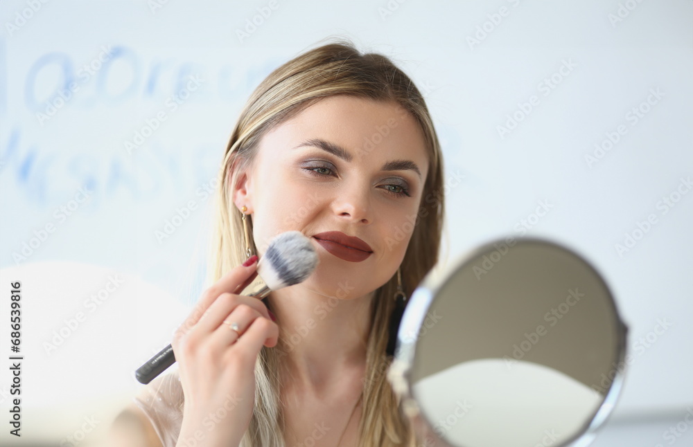 Smiling young woman looks in the mirror and paints herself with blush brushes. Makeup artist girl doing makeup learning with beauty and cosmetics video tutorials Smiling young woman looks in the mirror and paints herself with blush brushes. Makeup artist girl doing makeup learning with beauty and cosmetics video tutorials