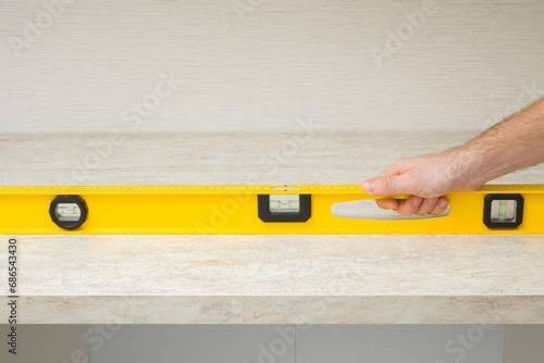 Young adult man hand using yellow spirit level and measuring horizontal surface of beige stone tabletop. Assembling new kitchen furniture. Closeup. Front view. Renovation process.
