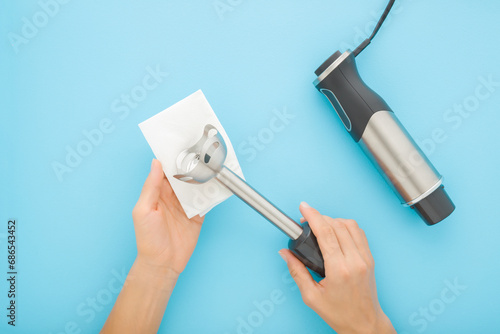 Foto Young adult woman hands cleaning immersion blender with dry white paper napkin on light blue table background