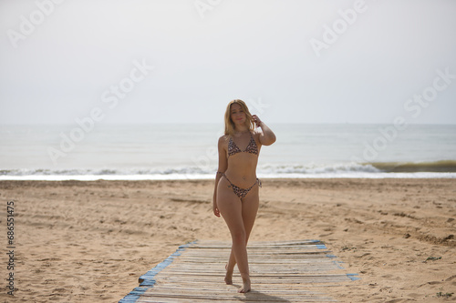 Young, blonde, beautiful woman, wearing a leopard print bikini, walking along a wooden walkway on the beach, smiling, happy, carefree. Concept beach, bikini, happiness, smile.