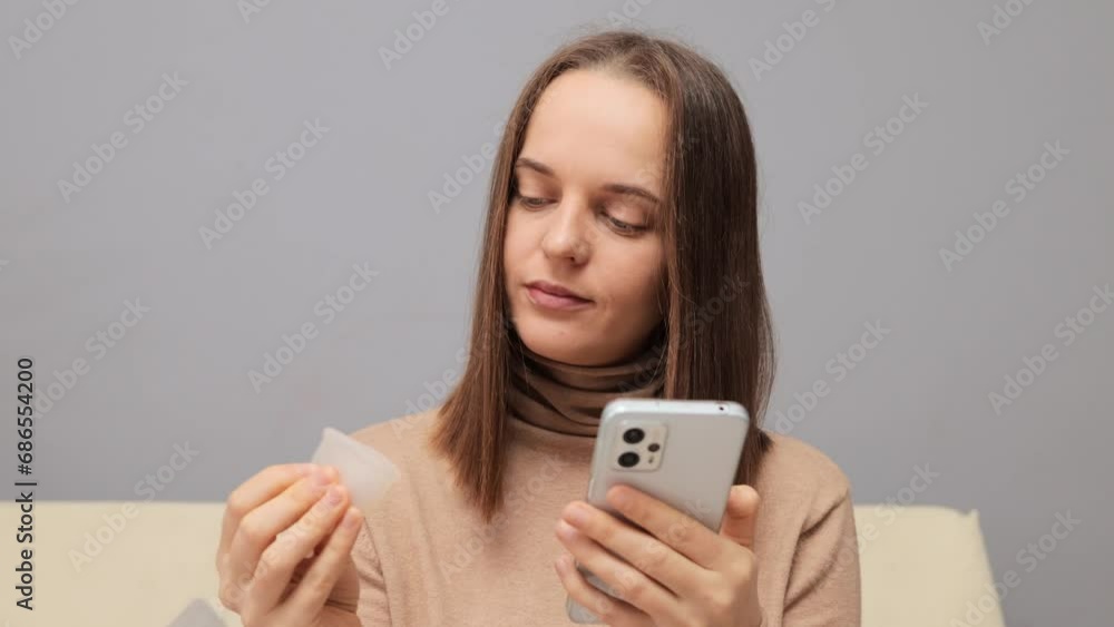 Concentrated adult young woman using mobile phone and holding reusable cup learning how to use alternative protection product searching tutoring video in internet. Concentrated adult young woman using mobile phone and holding reusable cup learning how to use alternative protection product searching tutoring video in internet.