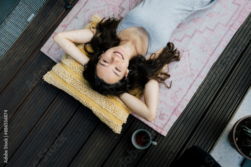 Directly above view of smiling woman lying down with hands behind head on rug in balcony