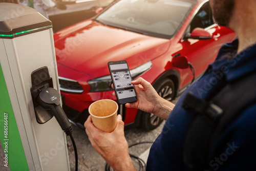 Man holding disposable cup using smart phone at electric car charging station photo