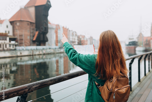 Attractive young female tourist is exploring new city. Redhead girl with backpack holding a paper map on city street in Gdansk. Traveling Europe in autumn. Famous Zuraw crane, Motlawa river photo